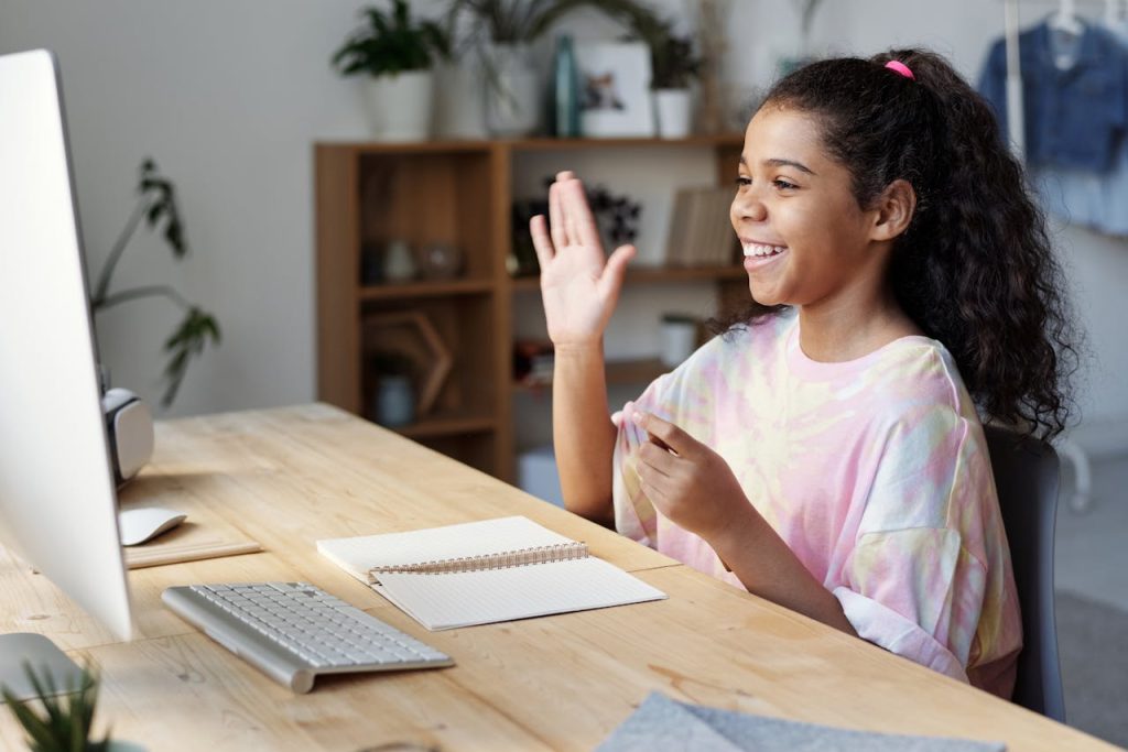 The Art of Drawing Readers In: Your attractive post title goes here A happy girl waves during an online class at home, sitting at a desk with a computer.