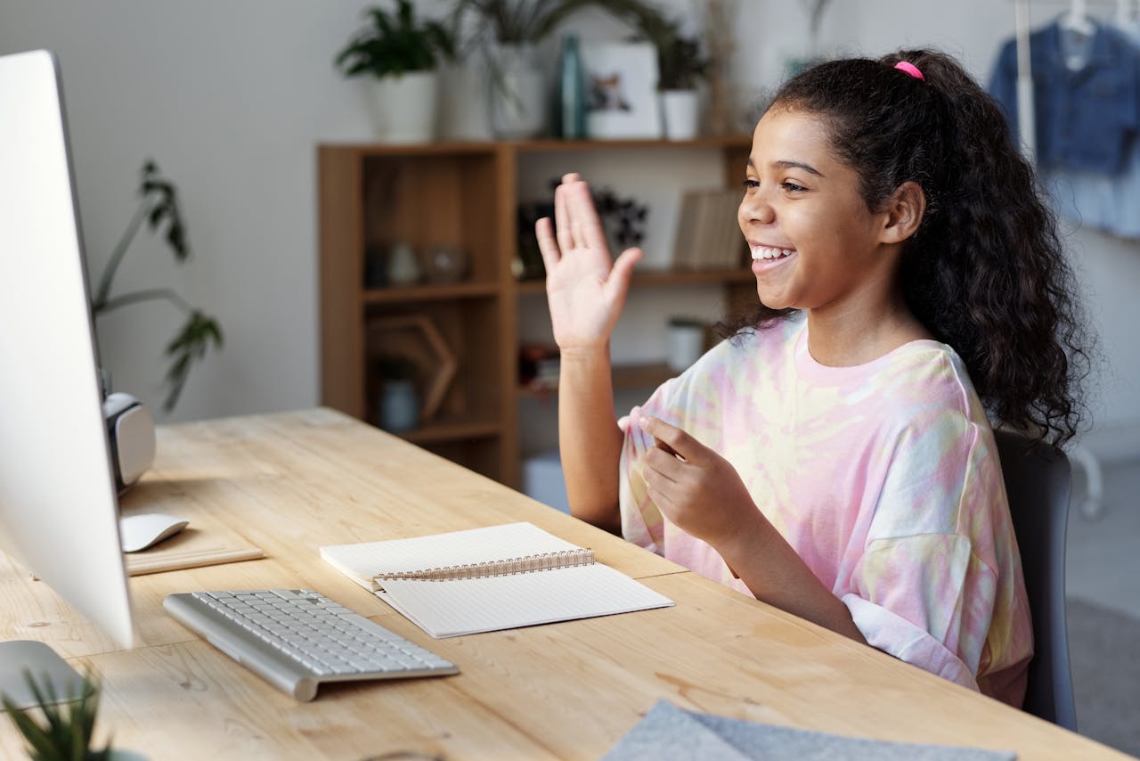 The Art of Drawing Readers In: Your attractive post title goes here A happy girl waves during an online class at home, sitting at a desk with a computer.